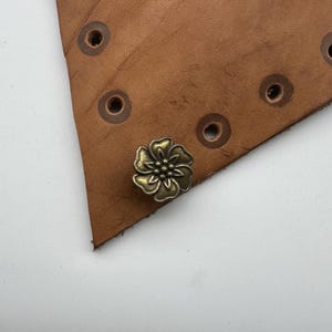 May include: A close-up of a gold-colored metal flower-shaped button. The button is attached to a piece of brown leather.