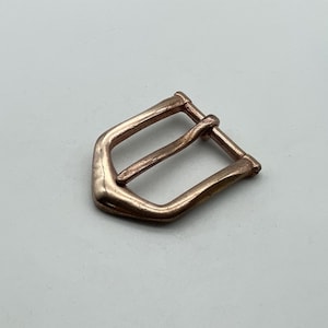May include: A bronze-colored belt buckle with a rectangular shape and a single bar. The buckle has a slightly curved design and a smooth finish. The metal buckle is a hardware component for belts.