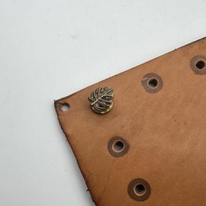 Op de afbeelding: Close-up van een bruine leren riem met verschillende gelijkmatig verdeelde gaten. Een decoratieve metalen blad-vormige stud is bevestigd aan het leer. Het leer heeft een natuurlijke, getextureerde uitstraling en de stud is bronskleurig.