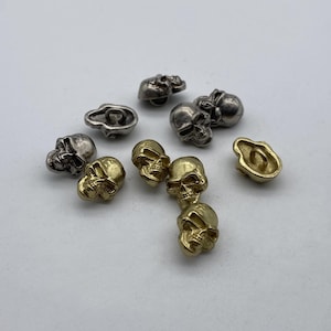 Könnte beinhalten: Eine Sammlung von 10 knopfartigen Schädeln in Silber- und Goldtönen. Die Knöpfe haben ein erhabenes Schädelmotiv und ein Loch in der Mitte zum Nähen.