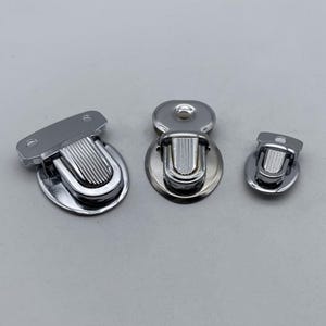 Pode incluir: Três fechos de metal prateados de diferentes tamanhos. Os fechos têm uma forma arredondada, ligeiramente abobadada, com um design nervurado em relevo no centro.