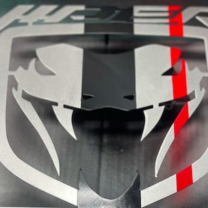 Puede incluir: Un emblema de coche de metal plateado con un diseño de serpiente en blanco y negro y una franja roja. La palabra "Viper" está por encima de la serpiente.