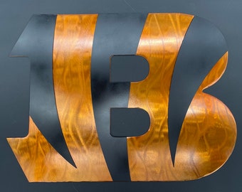 Pode incluir: Arte de parede em metal com a letra "B" com fundo preto e listras cor de cobre. O design tem uma textura de metal escovado, criando um visual moderno e industrial. Adequado para decoração de casa.