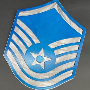 Pode incluir: Um emblema de metal azul e prateado com um design de estrela e listras. O emblema tem um fundo azul com listras prateadas e uma estrela no centro. Provavelmente uma insígnia de patente militar.