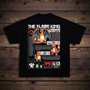 May include: Black t-shirt featuring the text "THE FLAME KING" above a graphic of Charizard, Charmeleon, and Charmander. The shirt includes information about the Pokémon characters. The shirt is on a wooden background.