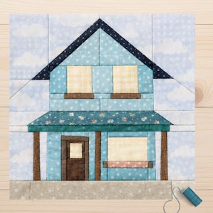 May include: A quilted fabric block depicting a house with a blue sky background. The house features a blue polka dot pattern, yellow window panes, and a floral print awning. Brown posts support the awning and a brown door is visible.