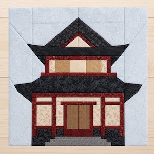 Op de afbeelding: Een vierkant stoffen quiltblok met een samengesteld ontwerp van een pagode met meerdere verdiepingen. Het gebouw gebruikt zwarte, donkerrode en beige stoffen met bloemen- en bamboepatronen, tegen een lichtblauwe achtergrond met witte bloesems.