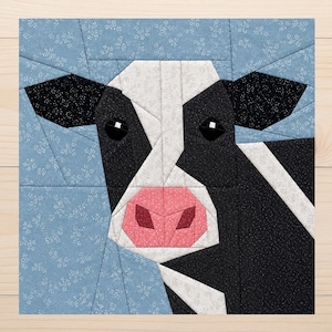 Pode incluir: Um bloco de patchwork de tecido com o rosto de uma vaca. A vaca tem marcas em preto e branco, um nariz rosa e olhos pretos. O fundo é azul claro com um padrão floral branco. O bloco quadrado provavelmente faz parte de um edredom ou projeto têxtil.