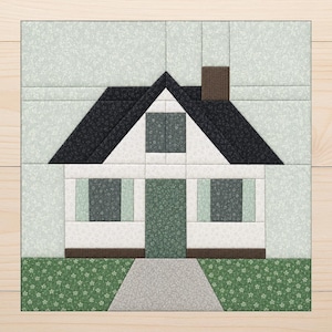 May include: A square fabric quilt block featuring a house design. The house has a dark charcoal roof, a brown chimney, white walls, and a green door. Two windows with green frames flank the door. A gray path leads to the house on green grass.