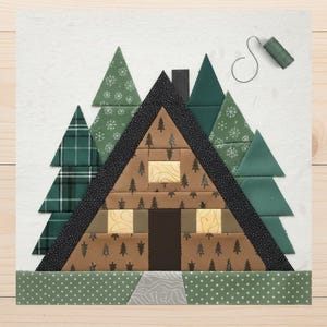 May include: A fabric block featuring a cabin design with green trees and a brown cabin with two yellow windows. The cabin has a black roof and chimney. The trees are various shades of green and patterned. A spool of green thread sits above the cabin.
