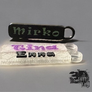 May include: Two black and white plastic keychains with the names "Mirko" and "Emma" printed on them. The keychain with the name "Mirko" is black and has a green font. The keychain with the name "Emma" is white and has a purple font.