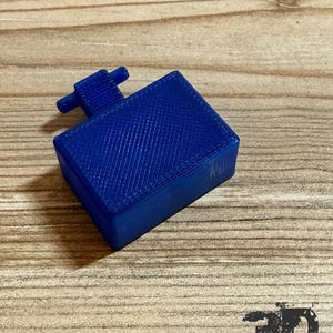 Op de afbeelding: Een rechthoekige, 3D-geprinte blauwe kunststof component met een getextureerde oppervlakteafwerking en een kleine, T-vormige uitstulping aan een van de zijden. Het object heeft een solide, doosachtige structuur met zichtbare lijnen van het printproces.