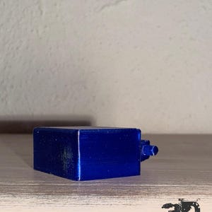 Op de afbeelding: Een klein, rechthoekig, 3D-geprint object in een levendige, metallic blauwe afwerking. Het item heeft een plat bovenoppervlak en een kleine, cilindrische uitstulping die aan &eacute;&eacute;n kant uitsteekt. De tekst "3D BlackMT" is zichtbaar in de rechterbenedenhoek.