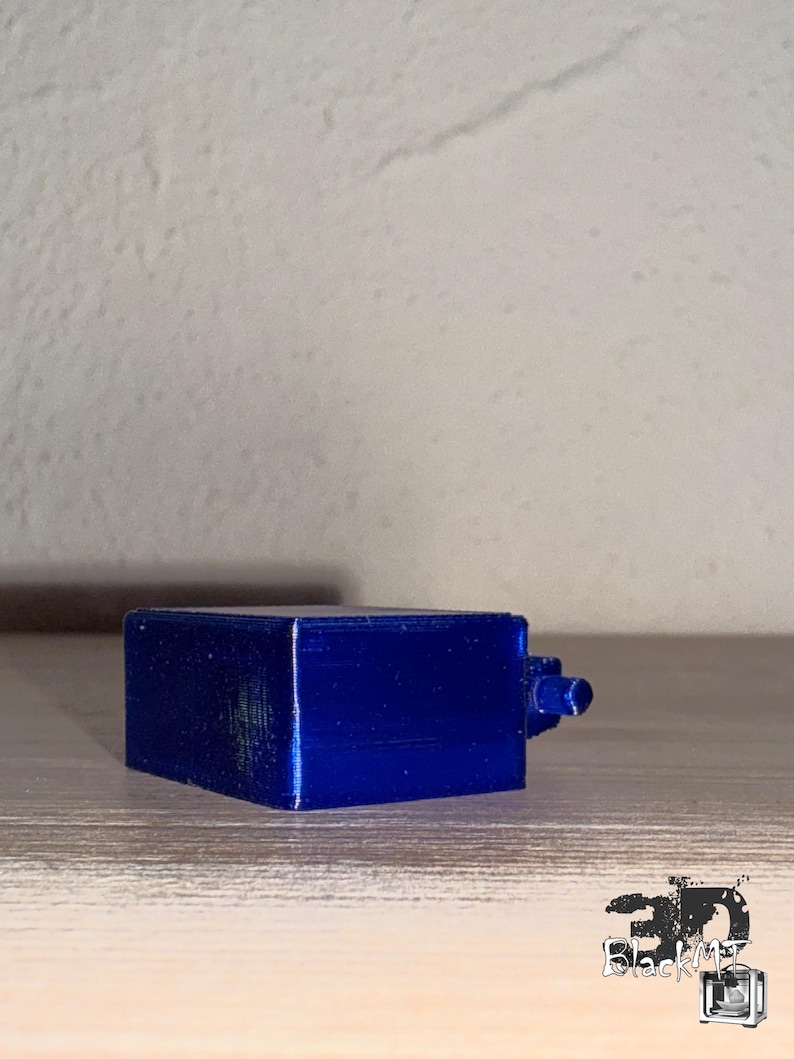 Op de afbeelding: Een klein, rechthoekig, 3D-geprint object in een levendige, metallic blauwe afwerking. Het item heeft een plat bovenoppervlak en een kleine, cilindrische uitstulping die aan &eacute;&eacute;n kant uitsteekt. De tekst "3D BlackMT" is zichtbaar in de rechterbenedenhoek.