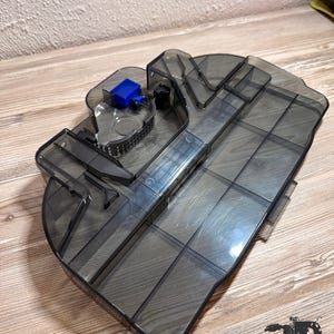 Op de afbeelding: Een doorschijnende donkergrijze plastic stofbak voor een robotstofzuiger. Het onderdeel is voorzien van een rechthoekig hoofdcompartiment met interne verdelers, een blauwe plastic vergrendeling en een zichtbaar gaasfiltergedeelte.