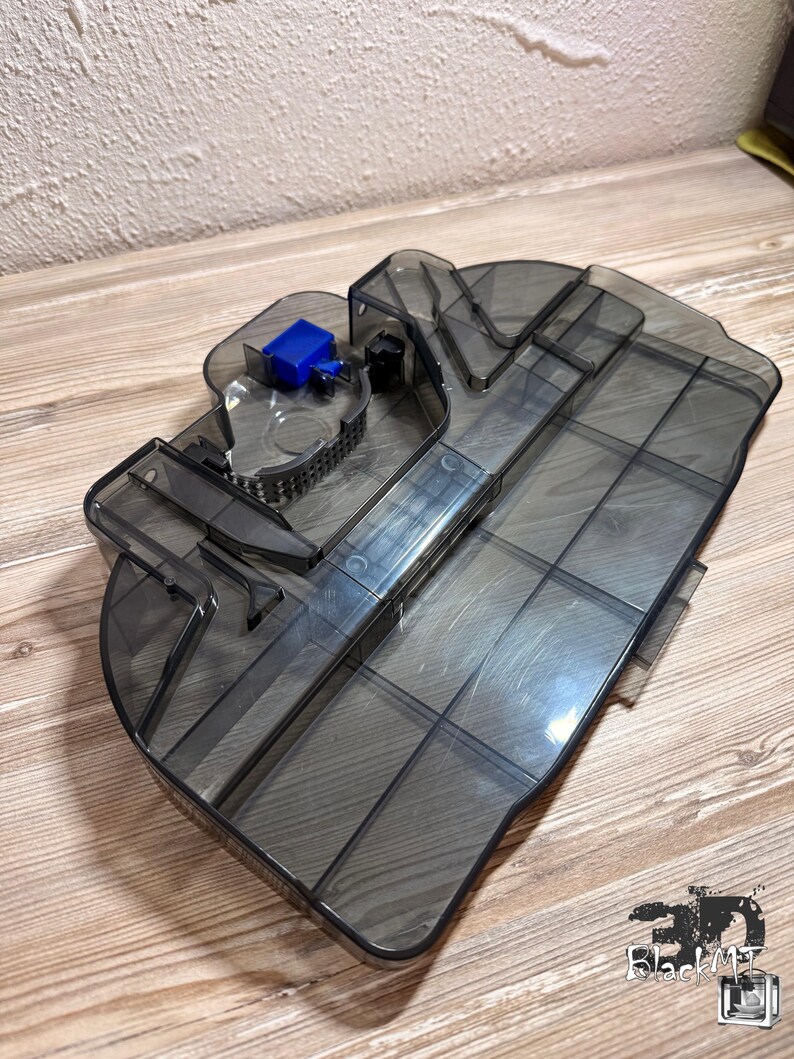 Op de afbeelding: Een doorschijnende donkergrijze plastic stofbak voor een robotstofzuiger. Het onderdeel is voorzien van een rechthoekig hoofdcompartiment met interne verdelers, een blauwe plastic vergrendeling en een zichtbaar gaasfiltergedeelte.