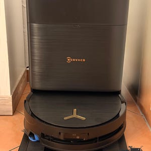 Op de afbeelding: Een donkergrijze Ecovacs robotstofzuiger die is aangemeerd bij zijn bijpassende rechthoekige oplaadstation. De stofzuiger heeft een cirkelvormig ontwerp met een goudkleurig embleem aan de bovenkant, terwijl het verticale station de merknaam Ecovacs in oranje weergeeft.