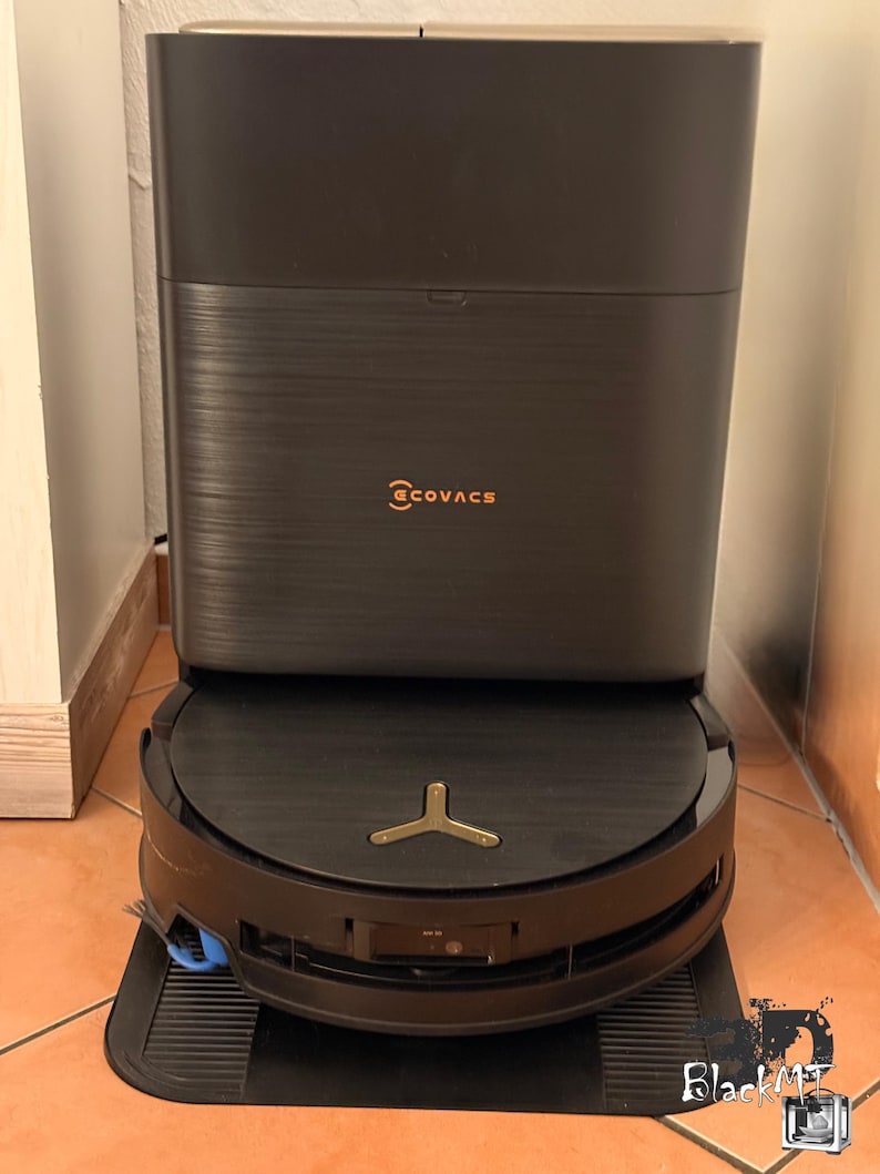 Op de afbeelding: Een donkergrijze Ecovacs robotstofzuiger die is aangemeerd bij zijn bijpassende rechthoekige oplaadstation. De stofzuiger heeft een cirkelvormig ontwerp met een goudkleurig embleem aan de bovenkant, terwijl het verticale station de merknaam Ecovacs in oranje weergeeft.