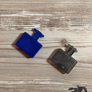 Op de afbeelding: Twee kleine rechthoekige kunststof onderdelen met uitstekende T-vormige lipjes. Het ene onderdeel is effen blauw met een getextureerd oppervlak, terwijl het andere doorschijnend donkergrijs is met een gladde afwerking en zichtbare details.