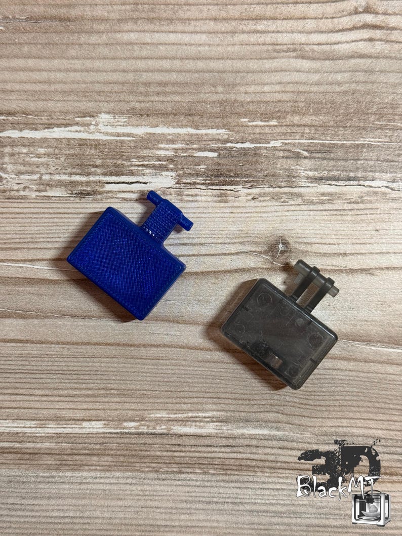 Op de afbeelding: Twee kleine rechthoekige kunststof onderdelen met uitstekende T-vormige lipjes. Het ene onderdeel is effen blauw met een getextureerd oppervlak, terwijl het andere doorschijnend donkergrijs is met een gladde afwerking en zichtbare details.