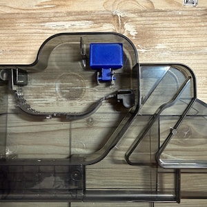 Op de afbeelding: Een doorschijnend donkergrijs plastic onderdeel met een complex intern railsysteem met opstaande randen en een enkel felblauw rechthoekig blok bevestigd aan een klein scharniermechanisme. De structuur bevat diverse kanalen en verdelers.