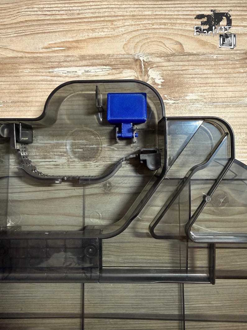 Op de afbeelding: Een doorschijnend donkergrijs plastic onderdeel met een complex intern railsysteem met opstaande randen en een enkel felblauw rechthoekig blok bevestigd aan een klein scharniermechanisme. De structuur bevat diverse kanalen en verdelers.