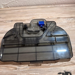 Op de afbeelding: Een doorschijnend donkergrijze plastic watertank voor een robotstofzuiger. Het onderdeel heeft een rechthoekige vorm met afgeronde randen, interne compartimenten, een kleine blauwe plastic motorbehuizing en een zwarte rubberen filterafdichting.