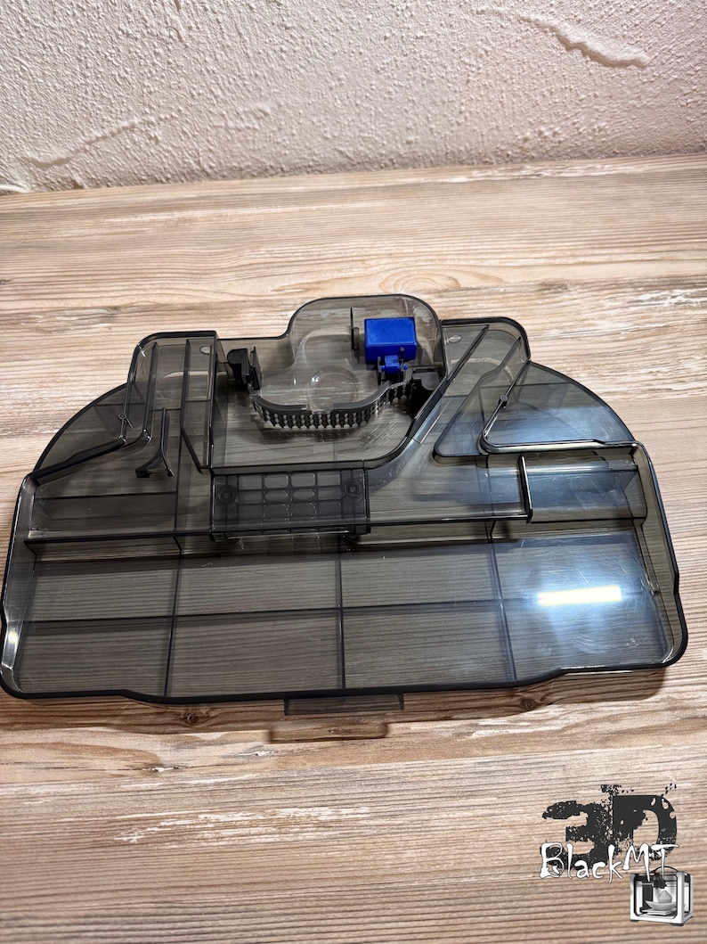 Op de afbeelding: Een doorschijnend donkergrijze plastic watertank voor een robotstofzuiger. Het onderdeel heeft een rechthoekige vorm met afgeronde randen, interne compartimenten, een kleine blauwe plastic motorbehuizing en een zwarte rubberen filterafdichting.