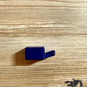 Op de afbeelding: Een kleine, massieve, donkerblauwe rechthoekige component met een enkele, dunne, uitstekende lip aan de rechterkant. Het object heeft een gladde, uniforme afwerking en een duidelijke geometrische vorm, die eruitziet als een enkel gegoten onderdeel.