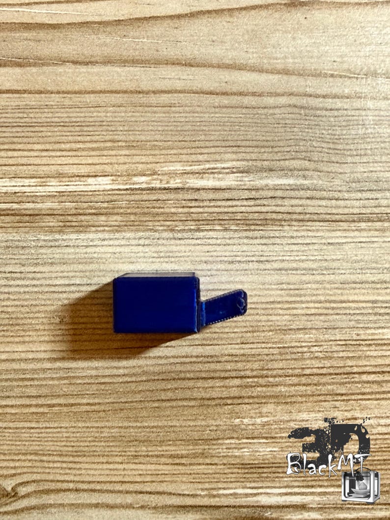 Op de afbeelding: Een kleine, massieve, donkerblauwe rechthoekige component met een enkele, dunne, uitstekende lip aan de rechterkant. Het object heeft een gladde, uniforme afwerking en een duidelijke geometrische vorm, die eruitziet als een enkel gegoten onderdeel.
