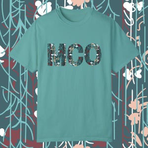 May include: A teal blue t-shirt with a floral pattern design that spells out the word "MCO" in large letters.