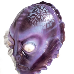 Peut inclure: Un masque d'extraterrestre violet avec un visage détaillé et de grands yeux blancs. Le masque a une surface texturée et un motif floral bleu clair sur le dessus de la tête. Le masque est probablement destiné au cosplay ou aux costumes.