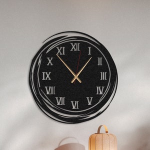 Może przedstawiać: Czarny zegar ścienny z rzymskimi cyframi i złotą wskazówką godzinową i minutową. Zegar jest zamontowany na białej ścianie nad drewnianym szafką z dwojgiem drzwi. Na szafce stoi lampa i książki.