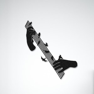 Op de afbeelding: Zwarte metalen wandkunst die twee handen toont die een muziekinstrument bespelen met noten die eruit vliegen. De kunst is gemonteerd op een witte muur in een slaapkamer.