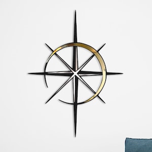 May include: A black metal wall decor with a gold circle in the center, resembling a compass rose. The decor is mounted on a white wall above a blue armchair with a white throw blanket.
