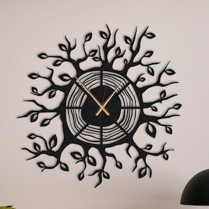 Puede incluir: Reloj de pared negro con diseño de rama de árbol. El reloj tiene agujas doradas y está colgado en una pared blanca sobre un escritorio de madera con una lámpara, un libro y un pequeño reloj que marca "SÁB 10".