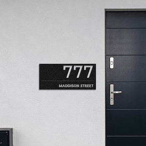 May include: A black and white house number sign with the number 777 and the street name Maddison Street. The sign is mounted on a white wall next to a black door.