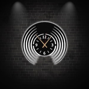 May include: A black and white clock with a circular design. The clock face has white numbers and hands. The clock is set against a dark brick wall.