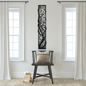 Puede incluir: Una pieza de arte mural de metal negro con un diseño de árbol con intrincadas ramas y hojas. La obra de arte rectangular está montada verticalmente en una pared blanca. Una silla de madera oscura con un cojín se encuentra debajo.