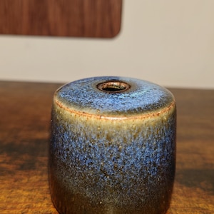 May include: A small, round, blue and brown ceramic vase with a hole in the top. The vase has a speckled glaze.