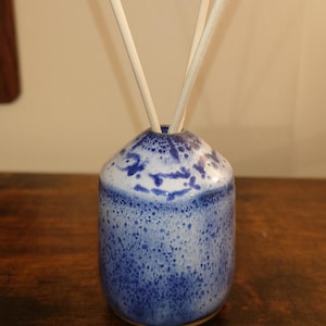 May include: A blue and white ceramic essential oil diffuser with a speckled design. The small, round vase holds several white reeds. The diffuser sits on a dark wooden surface. Home decor.