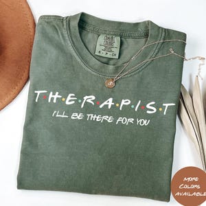 Op de afbeelding: Groen T-shirt met de tekst "THERAPIST I'LL BE THERE FOR YOU" in wit met gekleurde stippen. De tekst is in de stijl van de show Friends.