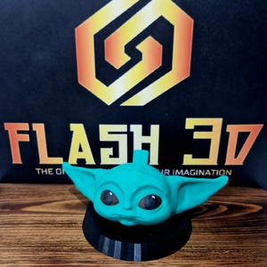 May include: A teal green 3D printed figurine of a character from the Star Wars franchise, known as "The Child" or "Baby Yoda". The figurine is sitting on a black circular base. The text "FLASH 3D" is visible in the background.