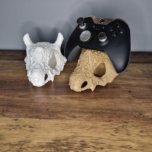May include: Two 3D printed skull-shaped stands, one white and one brown, with intricate designs. A black Xbox controller is resting on the brown skull stand.