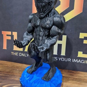 Puede incluir: Una figura negra de Venom impresa en 3D. La figura tiene un físico musculoso y detallado, y una cara amenazante con dientes afilados. Se encuentra sobre una base azul con la palabra "Venom" impresa. El fondo incluye el texto "THE ONLY" y "'S YOUR IMAGIN".