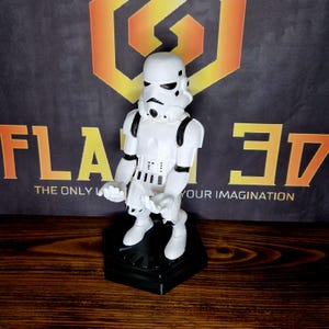May include: White plastic figurine of a Star Wars Stormtrooper with a black base. The figurine is posed in a standing position with its arms raised.