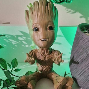 May include: A brown figurine of Groot from Guardians of the Galaxy. The figure has a smiling face, outstretched arms, and is seated. The figurine is surrounded by green foliage and a gray pot.