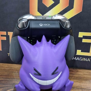 Op de afbeelding: Een paars Gengar Pokémon-figuur met een brede grijns, die een Xbox-controller in zijn handen houdt.