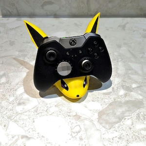 May include: A black Xbox controller rests on a yellow Pikachu-themed stand. The stand features Pikachu's face and ears, with the controller's buttons and joysticks visible. The controller is black with silver accents.