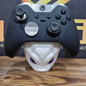 May include: A black and silver Xbox controller with a white faceplate and a white stand with purple eyes. The controller is resting on the stand.